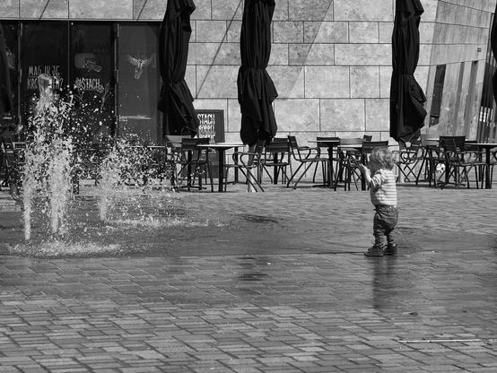 A young child stands playfully near a water fountain in a plaza, surrounded by outdoor seating with umbrellas. The image is in black and white, emphasizing the child's interaction with the water.