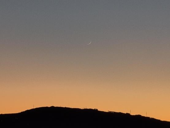 Fotografia após o pôr do sol, com a lua nova como um finíssimo crescente branco sobre o alaranjado do céu. Na parte inferior, o contorno negro de uma montanha.