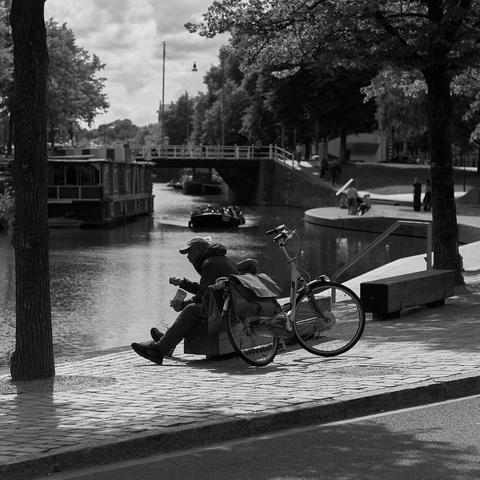 A person sits on the edge of a canal, playing a guitar and enjoying a snack. A bicycle rests nearby, and there are trees and a bridge in the background. The scene is depicted in black and white, capturing a serene moment by the water