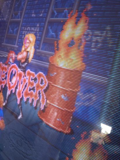 Dettaglio di una parte dello schermo CRT, con un barile di metallo rosso da cui escono delle fiamme, la parte finale della scritta "Game Over" e lo sprite di una donna bionda armata di frusta.