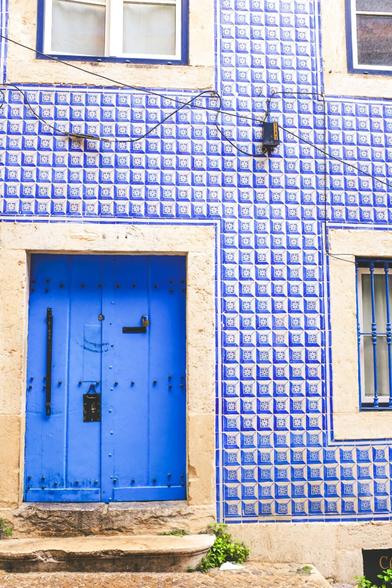 A vibrant street view of a traditional Portuguese building facade, featuring a bold blue wooden door framed by beige stone. The exterior wall is covered with classic blue and white azulejo tiles arranged in a repeating geometric pattern. A small step leads up to the door, and a patch of greenery grows at the base of the wall. Above the door are two windows with white curtains, also bordered by beige stone and blue trim. Black electrical wires cross the tiled surface, adding to the urban charm.