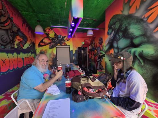 Paul, Abraxas3d, and Rose sit at a table at Arcade Monsters in San Diego. The walls are very brightly decorated with florescent paint. Table is garishly colored. After gaveling in, we voted on #DEFCON participation (unanimous yes), talked about the AMSAT-DL workshop,and then had a work session for #OpulentVoice.