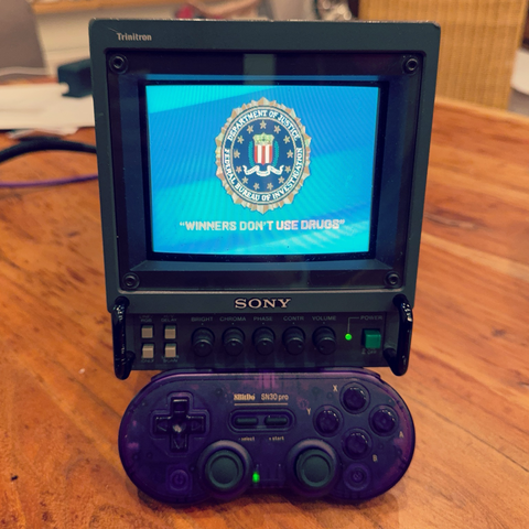 Das Bild zeigt ein kleines, vintage Sony Trinitron-Handheld-Gaminggerät, das auf einem Holztisch steht. Das Gerät hat einen kleinen Bildschirm, auf dem das Emblem des FBI und die Aufschrift "WINNERS DON'T USE DRUGS" zu sehen sind. Unter dem Bildschirm befinden sich verschiedene Steuerungen wie Bright, Chroma, Phase, Contrast und Volume. Vor dem Gerät liegt ein transparentes, violettes SBitDo SN30 Pro-Controller. Der Hintergrund ist unscharf, aber man kann einen Teil eines Raumes erkennen, der möglicherweise ein Wohnraum oder ein Arbeitsbereich ist. Bereitgestellt von @altbot, privat und lokal generiert mit Ovis2-8B 🌱 Energieverbrauch: 0.212 Wh