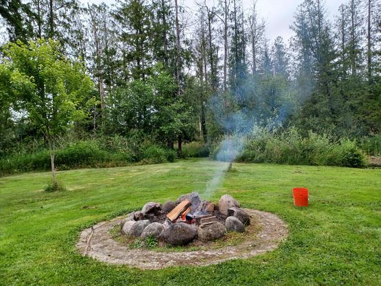 A natural stone fire pit with cut firewood burning and smoking surrounded by green grass against a stand of green shrubs and trees in the background. There is an orange 5 gallon bucket on the right for fire safety.