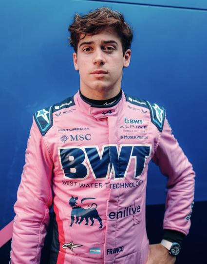 Formula 1 driver Franco Colapinto in a pink racing ensemble, standing in front of a blue wall. Black letters across his chest read BWT. His left hand rests on his hip.
