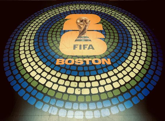 A bold and vibrant circular design featuring the FIFA World Cup trophy. Radiating outward from the center are concentric rings made up of rounded squares in shades of blue, green, and white, giving the design a modern, digital mosaic feel. At the heart of the circle is a prominent “26” in bright orange, with the trophy positioned squarely between the numbers. Just beneath it, “FIFA” appears in thick black lettering, and “BOSTON” sits in matching orange below, clearly indicating the city’s role as a host for the 2026 FIFA World Cup.