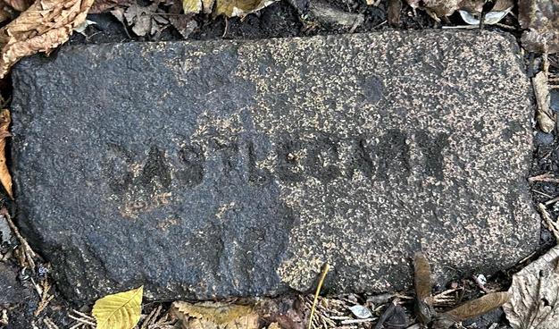 Surrounded by dead leaves, an old sandy-coloured but mud-covered clay brick is lying deeply embedded in the ground. The brick bears the stamp of the manufacturer - CASTLECARY