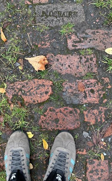 The viewer is looking down at the photographer's shoes on the ground and beneath them is a very old brick-paved walkway. Surrounded by dirt and dead leaves, most of the bricks are reddish coloured although there is one sandy-coloured but mud-covered brick bearing the stamp of the manufacturer - BLOCHAIRN