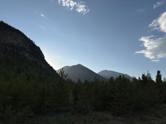 A scenic view of mountains silhouetted against a blue sky, with light peeking through. The foreground features pine trees and a blend of greenery, creating a serene natural landscape.