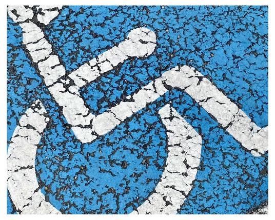 Close-up view of a weathered blue painted surface with a white wheelchair symbol, showing cracked paint and the underlying asphalt texture.