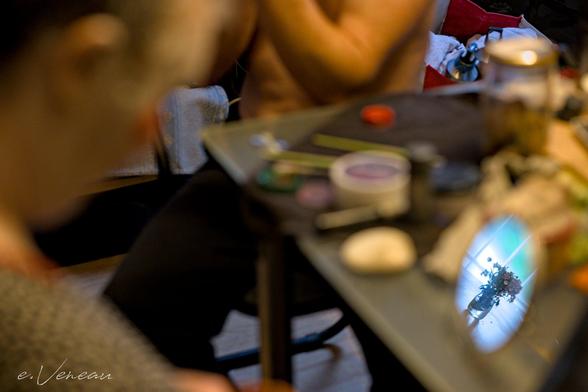 Blurred photograph of a black table with make-up in disarray. We can make out the two bodies partially in the image of the two artists applying make-up in their dressing room. The only thing in focus, in a round mirror on the table: a bouquet of wild flowers.