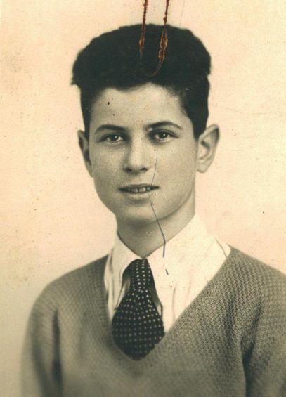 Portrait of a young boy wearing a v-neck sweater over a collared shirt, paired with a patterned tie. The boy has short, wavy hair and is looking directly at the camera, giving a subtle, pleasant expression.