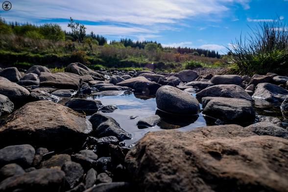 Low level shot of stones in river. Trees and moorland in background under blue cloud streaked sky