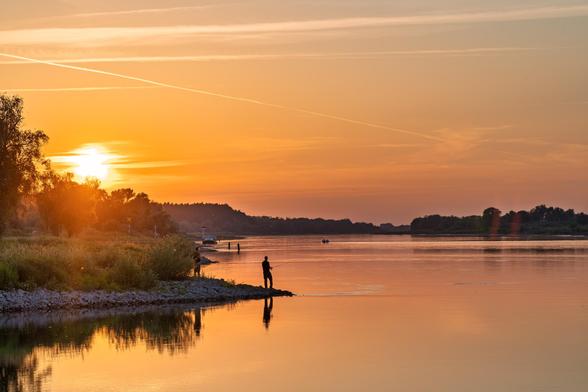 Blick entlang des Flusses. Das Ufer auf der linken Seite. Eine Silhouette eines Anglers ist am Ufer zu sehen im Hintergrund Bäume und die untergehende Sonne. Gegenlicht, das Wasser ist orange und die Landschaft Silhouettenhaft.