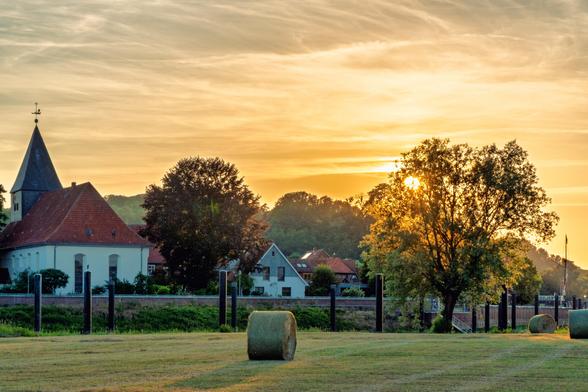Blick über die Wiesen. Vorne ein Heu allen.
Dahinter die stadt.links weiße Kirche mit rotem Dach und grauer Turmspitze. Rechts die untergehende Sonne hinter einem Baum.