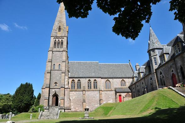 The Cathedral of the Isles in Millport. The image shows a view up and across grassy banks to a small stone church with a very tall tower and spire on its left. There is a range of further buildings on the right, at the top of a grassy bank. The sky is largely blue.