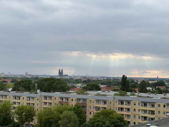 Blick über die Dächer von Magdeburg, ihm Vordergrund sind Wohnblöcke mit Balkonen, zwischendrin Bäume und am Horitont der Magdeburger Dom zu sehen, Der Himmel ist bewölkt und aus einer kleinen Lücke heraus sind einige Sonnenstrahlen erkennbar