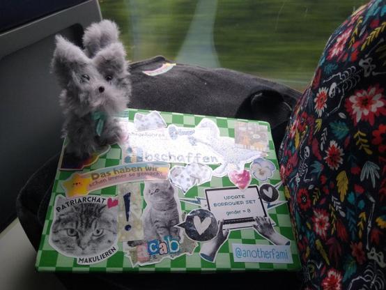 Foto einer Ablage im Regionalexpress. Im Hintergrund ist zu erkennen, dass der Zug fährt. Ein Pappschild liegt auf einer Umhängetasche rechts in Bild ist der Beutel in dem ich das Bastelmaterial transportiere zu sehen. Auf dem Pappschild klebt eine Collage aus den Stickern, die  ich zur BiblioCon mitgebracht habe. Darunter unter anderem der Publikumsliebling "Patriarchat makulieren" und "Das haben wir schon immer so gemacht". Rechts oben auf dem Pappschild sitzt eine graue Katze aus Pfeifenputzern. Sie trägt eine kleine Schleife und guckt hoffnungsvoll.