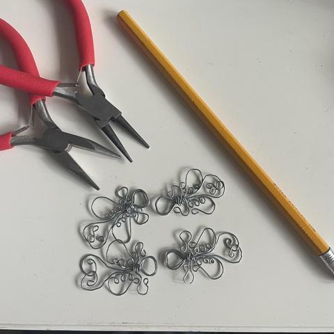 Two pairs of pliers with red handles in the top left corner, an unsharpened hb pencil to the right, and four little wire wrapped clasps in the lower middle. The clasps have decorative bulbs, reminiscent of a rococo or art noveau style