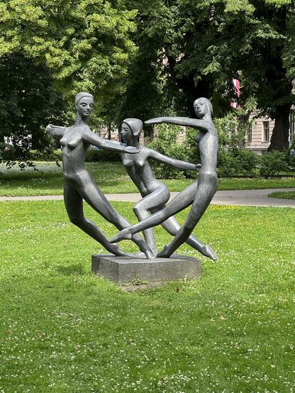 Eine Bronzeskulptur "Tänzerinnen", die drei stilisierte weibliche Figuren in Bewegung darstellt, auf einem Steinsockel. Die Figuren sind in einer dynamischen Pose angeordnet, wobei eine Figur die Hand ausstreckt, während die anderen eng interagieren. Die Skulptur ist umgeben von üppigem grünem Gras und Bäumen.