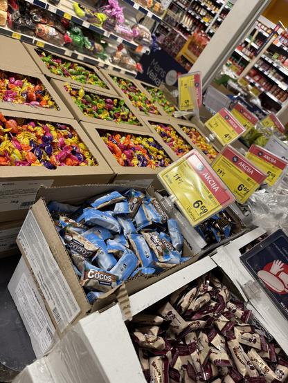Eine Auswahl an bunten Süßigkeiten in offenen Schachteln in einem Supermarkt, darunter in Folie eingewickelte Süßigkeiten, ist prominent in einem Geschäft ausgestellt, zusammen mit Preisschildern mit Rabatten. Lose Bonbons, wie in meiner Kindheit.