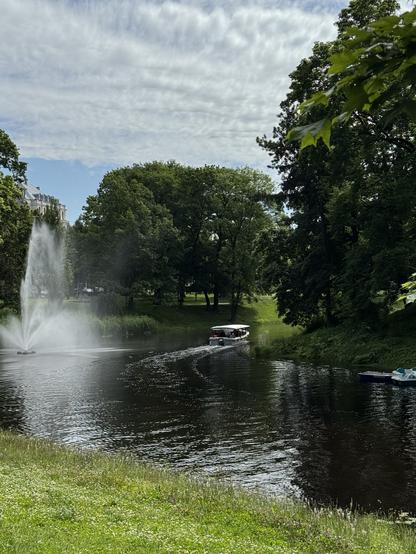 Eine ruhige Parkszene mit einem Kanal mit einem Brunnen, umgeben von üppig grünen Bäumen. Ein kleines Boot hinterlässt eine Spur, während es durch das Wasser navigiert, mit einem Blick auf Tretboote in der Nähe. Der Himmel ist teilweise bewölkt, was zur ruhigen Atmosphäre beiträgt.