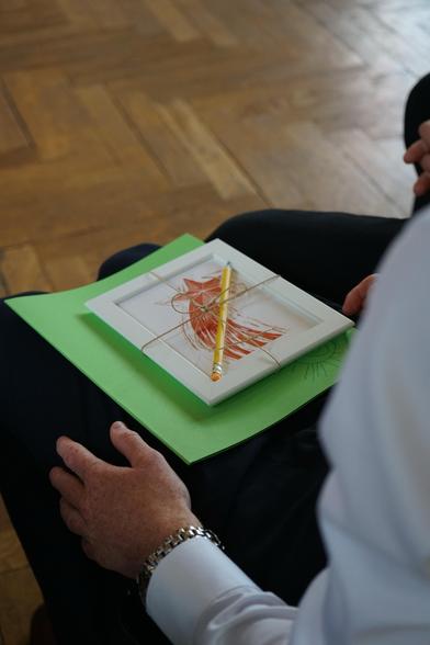 Das Bild zeigt auf dem Schoß einer Person liegend eine grüne Zeugnismappe und ein gerahmtes Bild mit Bleistift, mit Schleife als Geschenk verpackt.