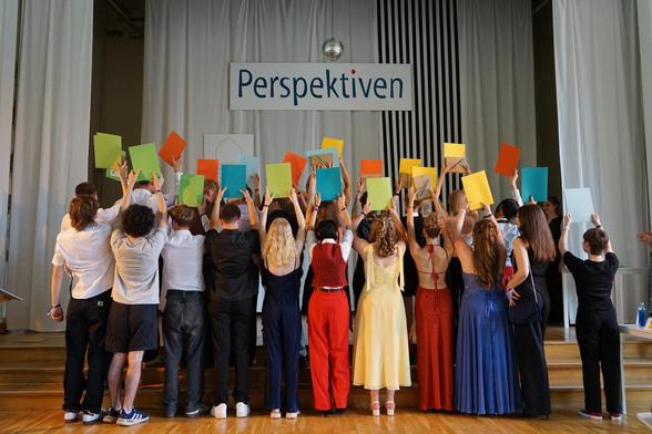 Das Bild zeigt eine Gruppe von festlich gekleideten Jugendlichen von hinten, die auf der Bühne unter einem großen Schild mit der Aufschrift "Perspektiven" ihre Zeugnisse hochhalten.