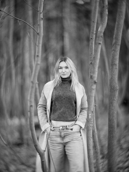 Woman in jeans and coat standing between two small trees in a forest. Swirly bokeh.