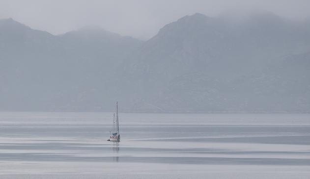 A single yacht on a still sea. Misty hills in the background.