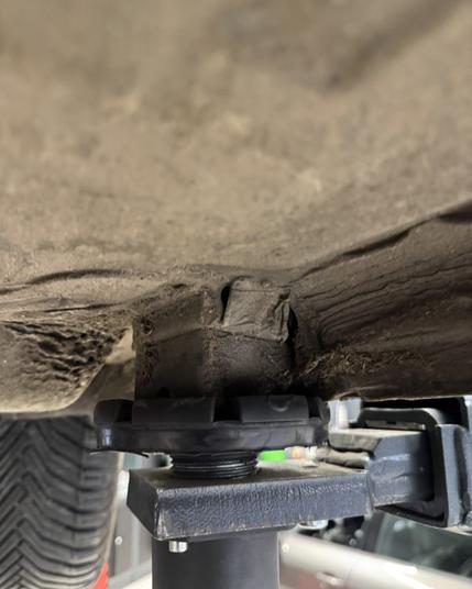 Ein schwarzer Hebebühnenstützpunkt hebt die Unterseite eines schmutzigen Fahrzeugs an, wobei sich ein Gummiklotz zwischen dem Stützpunkt und dem Fahrzeugrahmen befindet.