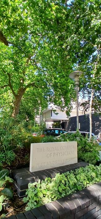 A scene from the front area of the Royal College of Physicians in London, England. There is a carved stone sign with the name and address of the building in the foreground and through mature trees and planting (dappled with sun and shade) lurks Dennis Lasdun's finest building, the front of which is a creamy ivory white block with two sections cantilevered above the ground.