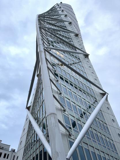 A modern, twisting skyscraper with a glass facade, viewed from below against a cloudy sky. The building features intricate steel supports and a distinctive spiral design.