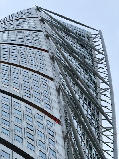 A close-up view of a modern, curved skyscraper with a distinctive design. The building features multiple windows and metal structural elements, including diagonal supports. The top of the building is partially obscured by the sky.