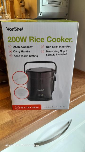 A box containing a mini rice cooker from Lidl UK. The box has the image of a small, black, cylindrical rice cooker.
The brand is VonShef.
The rest of the writing says:
200W Rice Cooker
300ml capacity
Carry handle
Keep warm setting
Non-stick inner pot
Measuring cup and spatula included
Dimensions: 18 x 16 x 15cm

The box is on my kitchen worktop with a white fronted drawer underneath.