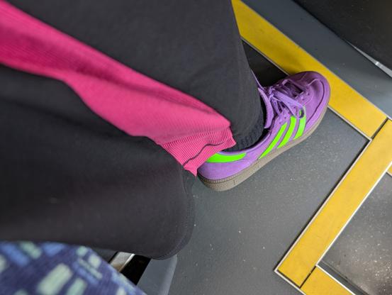 My right leg stretched out to show my purple and neon green trainers and black and hot pink trousers. My leg is partially stretched across the yellow caution strips on the bus floor.