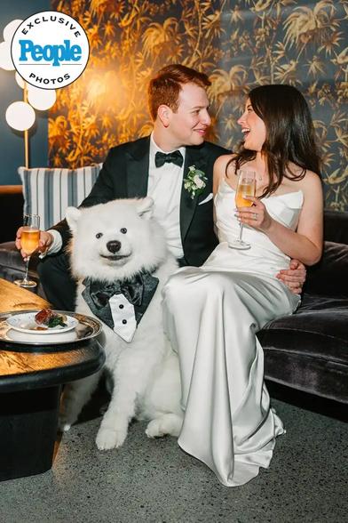 A dog wears a tuxedo costume next to a bride and groom at their wedding reception