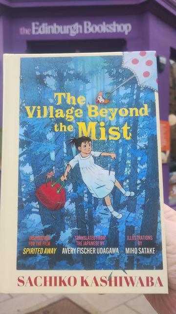 A copy of The Village Beyond the Mist, showing a young girl floating in a forest, held up outside the Edinburgh Bookshop