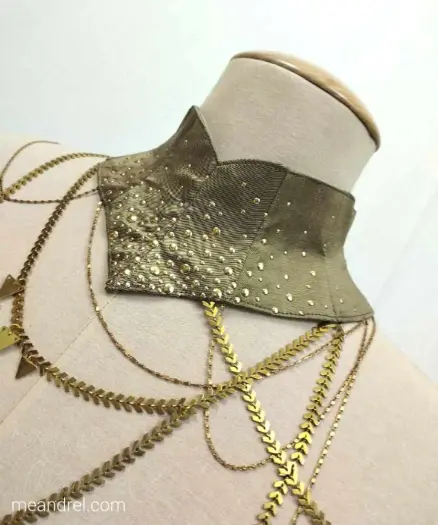 Photo d'un collier textile sur mannequin ; il est très large, doré, avec un dégradé de petits points métalliques dorés et de nombreuses chaines or qui retombent sur le torse !