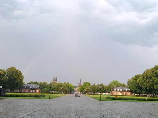 Foto vom Schlossplatz in Münster vom Schloss aus gesehen. Im Vordergrund ist eine mit Kopfsteinpflaster gepflasterte Fläche. Ein Weg mit dem gleiche Pflaster geht in der Bildmitte in den Hintergeund. Links und rechts daneben sind Grünflächen mit kleinen Nebengebäuden. Das rechte Häusschen glüht fast, weil es von der Sonne beschienen wird. Der Himmel sieht nach Regen aus. Links über dem Nebengebäude startet ein Regenbogen, der aber aber der Mitte des Bildes nicht mehr zu sehen ist. Ganz im Hintergrund und an den seitlichen Bildrändern sind Bäume und noch weiter im Hintergrund die Kirchtürme der Überwasserkirche, vom Dom und der Lambertikirche zu sehen.