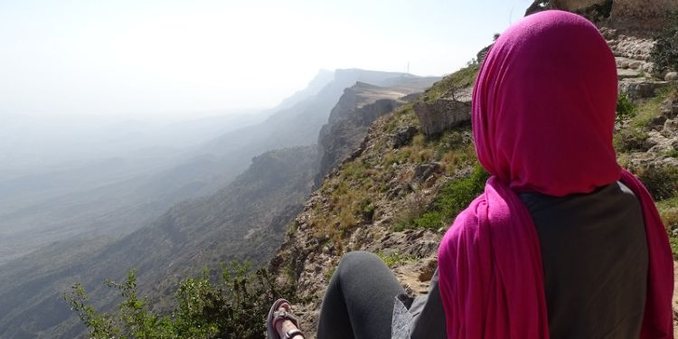 Figure in a red hijab looking out over misty, verdant cliffs in what looks to be a South Arabian landscape