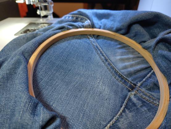 The crotch area of a pair of blue jeans in an embroidery hoop, a sweing machine in the background. There's a lighter and worn out area of fabric that's been sewn over in a grid-like pattern. There was a hole in the fabric there, only barely visible now. The idea is to continue filling the worn out area with stitches to repair it.