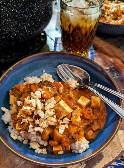 Dunkler Tisch, darauf ein blauer, tiefer Teller, darin Reis, Gemüsecurry mit Tofuwürfeln in Tomatensauce, Darübergestreut als Topping gehackte Cashews. Im Teller liegen auerßdem eine Gabel und ein Löffel. Im Hintergrund zu sehen: Kochtopf, Glas mit Eistee und Eiswürfeln, weiterer, gefüllter Teller.