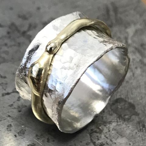 A close-up of a unique ring featuring a textured silver band with a smooth, gold snake-like design wrapped around it. The background is a subtle, blurred metallic surface.