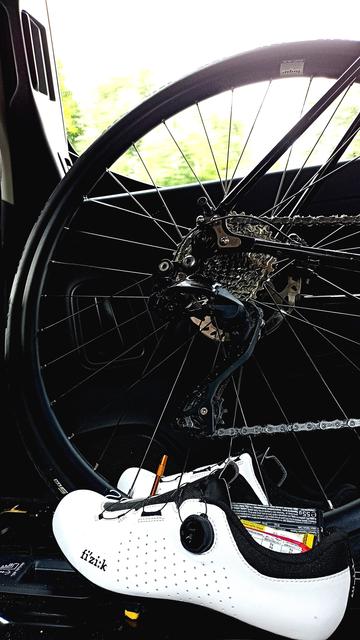 Ein Hinterrad von einem Rennrad in einem Auto. Im Vordergrund ein Paar weiße Radschuhe. In der Radschuhen sind Riegel und Gels verstaut.