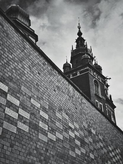 A low-angle, black and white shot of a brick wall on the left, adorned with numerous rectangular plaques inscribed with text. The wall recedes into the background, leading the eye towards a grand, ornate building with a prominent spire and multiple turrets, visible on the right side of the frame under a cloudy sky.