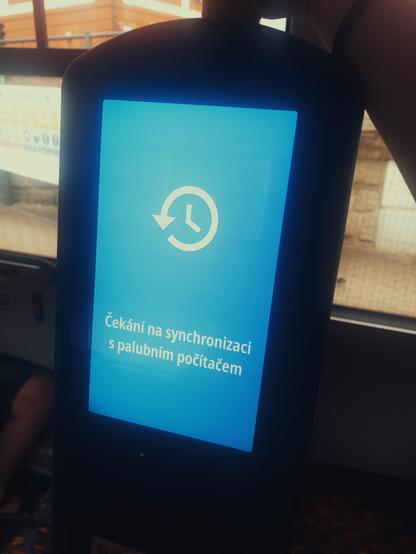Na obrázku je čelní pohled na stojan s modrým displejem odbavovacího automatu v tramvaji, zobrazující bílou ikonu hodin se šipkou v kruhu a text „Čekání na synchronizaci s palubním počítačem“.
