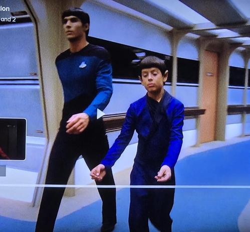 A Vulcan child and his father hurry down an Enterprise hallway. They are not carrying anything. They are both dressed in blue and black although the father is in uniform.