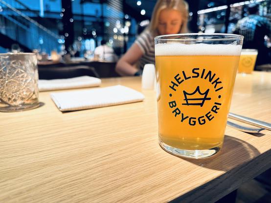 A glass of Helsinki Bryggeri IPA sits on a light wooden table in a hotel restaurant in Helsinki. The beer has a hazy golden color with a frothy white head, and the glass is branded with the Helsinki Bryggeri logo. The background features a softly lit, modern dining area with other patrons and tables, creating a relaxed and inviting atmosphere.