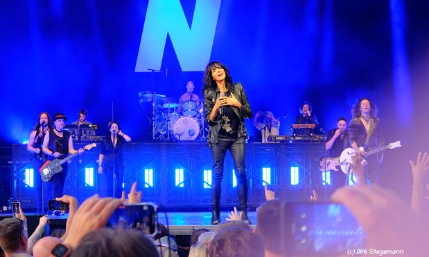 „Nena steht in schwarzer Lederkleidung im blauen Bühnenlicht auf der Bühne. Ihre rechte Hand hält ein Mikrofon, die linke liegt auf ihrer Brust. Sie blickt dankbar ins Publikum. Um sie herum die gesamte Band. Mehrere Menschen im Publikum fotografieren mit ihren Handys.“